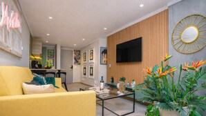 A modern living room with a yellow sofa, a glass coffee table, a flat-screen TV, wooden paneling, and a large potted plant with tropical flowers. - Ayamonte - 301b in Medell n (Medellín)