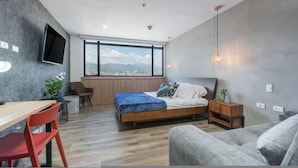 Un dormitorio moderno con cama, escritorio, silla, sofá, televisor montado en la pared y un ventanal grande.