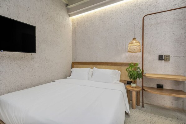 A bedroom with a bed, a wall-mounted TV, a hanging light fixture, a bedside plant, and wooden shelves. - Botánica Básica (Medellín)