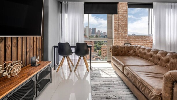 A modern living room with a leather sofa, a TV mounted on a wooden panel wall, a dining table with chairs, and a large window with a city view. - Oslo 2101 in Medell n (Medellín)