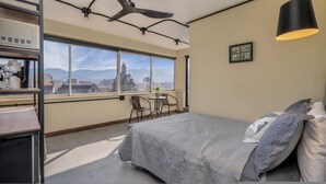 Un dormitorio con cama, escritorio, silla, ventana con vista a la ciudad y ventilador de techo.