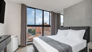 A modern hotel room with a large bed, a flat-screen TV, a desk, and a view of the cityscape through large windows. - Housy Laureles - 401 (Medellín)