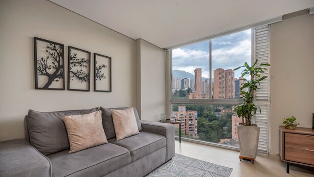 Ein modernes Wohnzimmer mit grauem Sofa, einem großen Fenster und Blick auf die Stadt sowie einer Zimmerpflanze.
