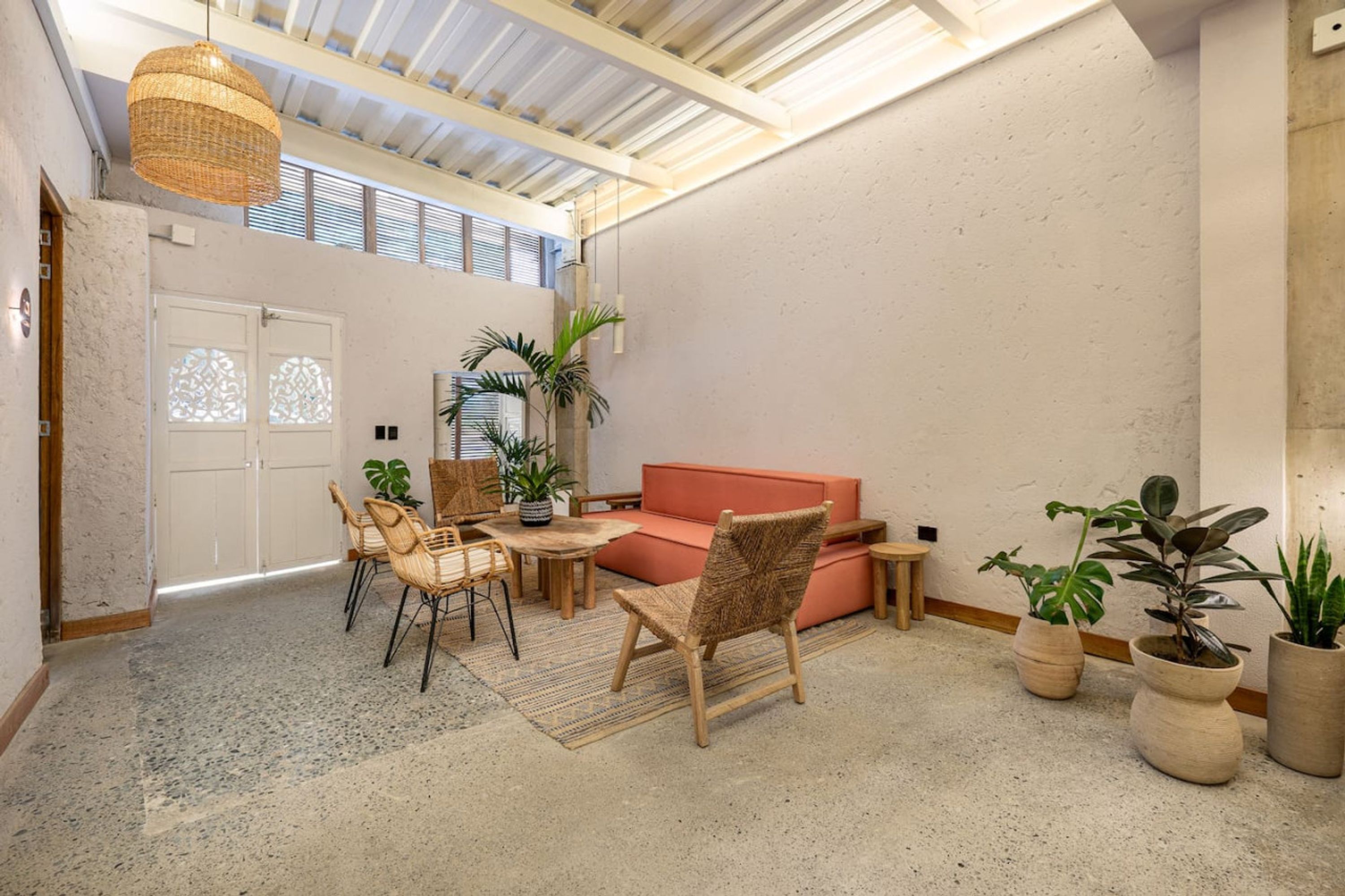 Une pièce spacieuse avec un plafond haut, un luminaire suspendu en fibres tressées, un canapé, une table à manger avec des chaises et plusieurs plantes en pot.