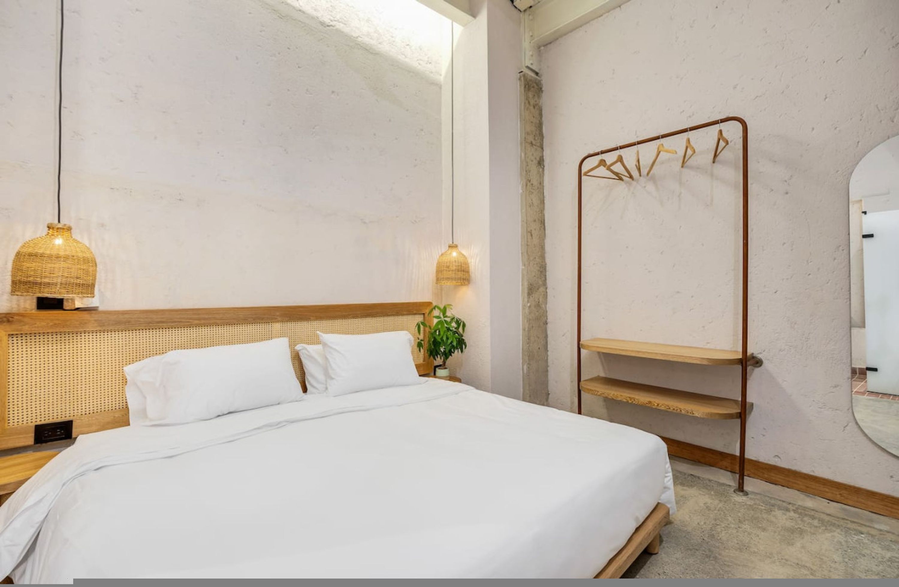 A bedroom with a large bed, two hanging lamps, a wooden headboard, a plant, a metal drying rack, and a mirror.