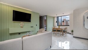 En moderne stue med et fladskærms-tv, en sofa, et spiseområde med bord og stole, og udsigt til bygninger gennem et stort vindue.