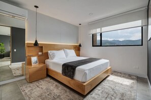 A modern bedroom with a wooden bed frame, bedside tables, a large window with a mountain view, and a hanging pendant light. - Go Living Suites - 508 (Medellín)