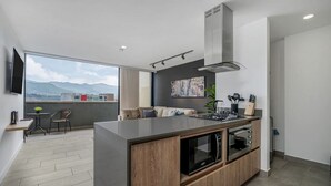 Een moderne keuken met een centraal eiland, een inbouwoven en een magnetron. Er is een eethoek met een tafel en stoelen, en een groot raam met uitzicht op de stad.