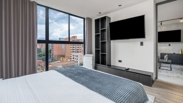 Una habitación de hotel moderna con una cama grande, un televisor de pantalla plana montado en la pared, un armario empotrado y vistas a la ciudad.