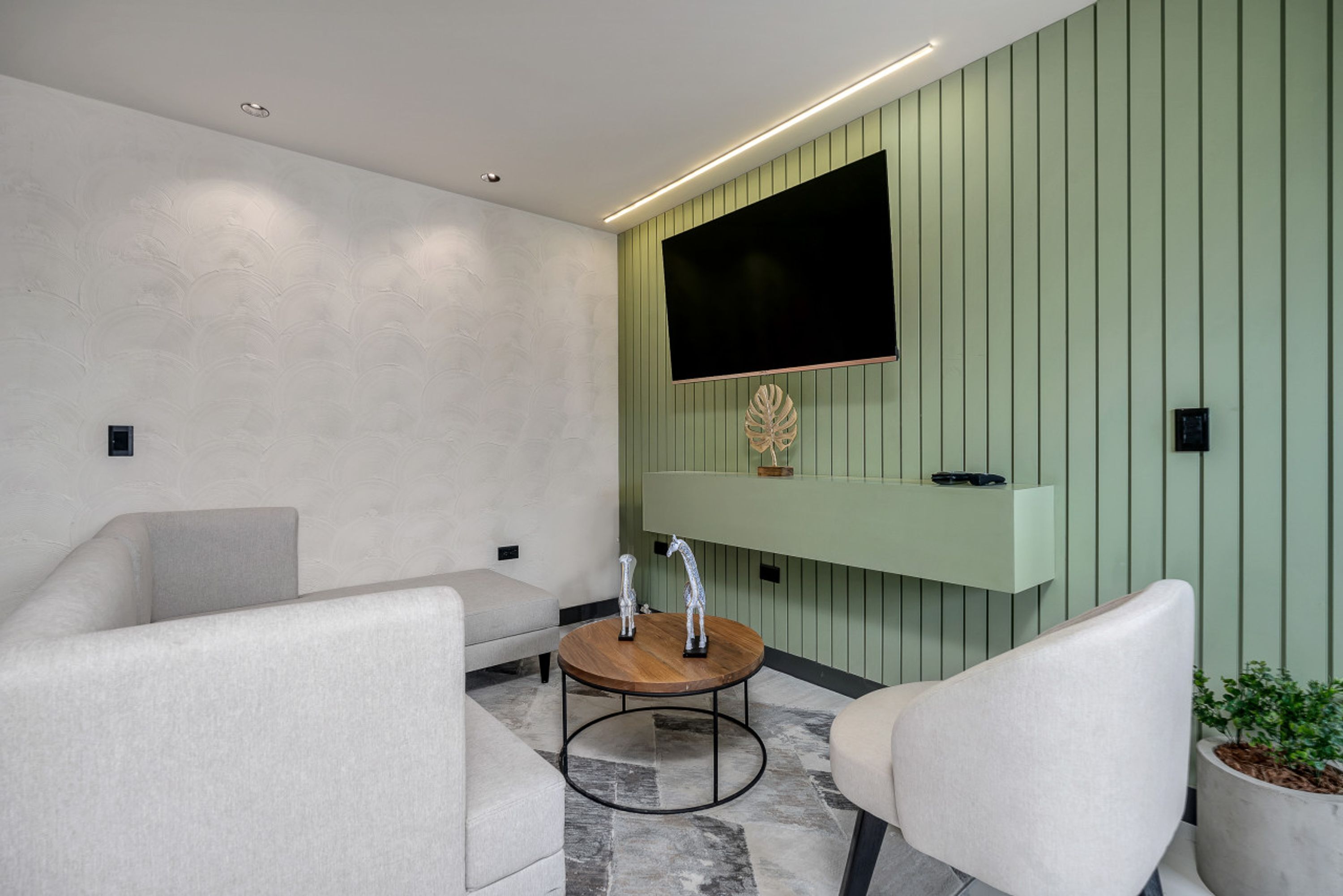 A modern room with a sofa, a round table with a coffee pot, a flat-screen TV mounted on a green wall, and a potted plant.