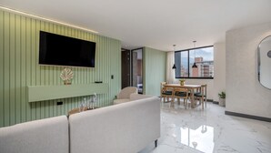 Un salon moderne doté d’un téléviseur à écran plat, de panneaux muraux verts, d’un canapé, d’un coin repas avec une table et des chaises, et d’une grande fenêtre offrant une vue sur la ville.