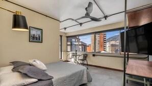 A bedroom with a bed, a desk, a chair, a ceiling fan, a window with a city view, and a framed picture on the wall. - Nutibara Express - 828 (Medellín)