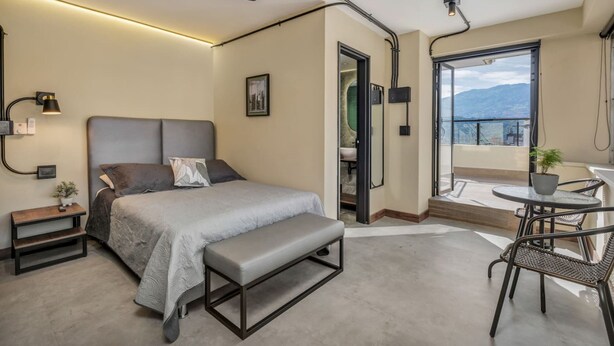 A modern bedroom with a bed, bench, bedside table, and a balcony with a table and chairs. - Nutibara Express - 828 (Medellín)