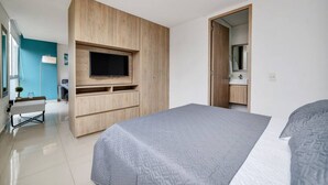 Une chambre d’hôtel moderne avec un grand lit, un téléviseur fixé sur un meuble en bois, un bureau avec une chaise et une salle de bain visible à travers une porte ouverte.