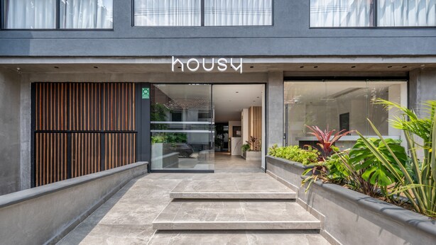 En modern entré till en byggnad med en skylt som visar ordet ”Housu”, en glasdörr och en betongtrappa som leder in.