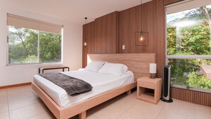 Een moderne slaapkamer met een houten hoofdbord, een groot raam met uitzicht op bomen, een nachtkastje met een lamp en een staande heater.