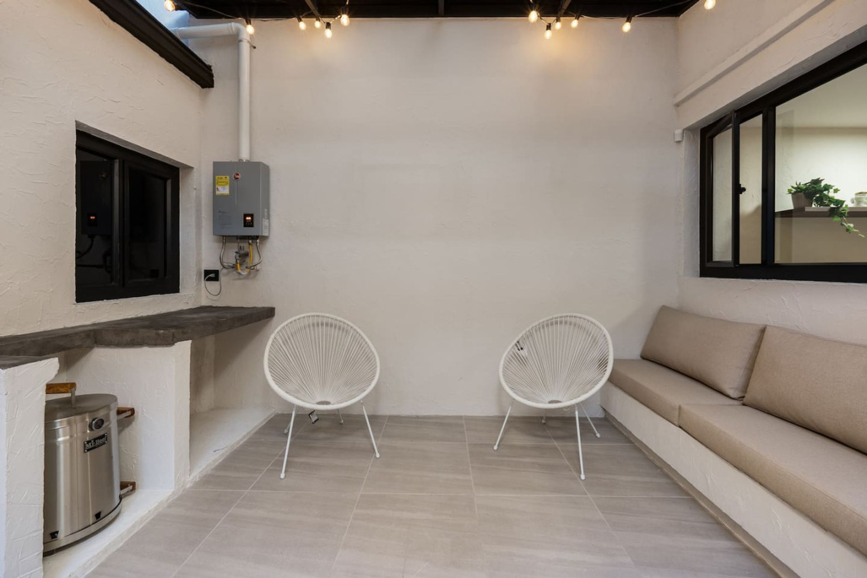 A room with a white wall, a beige sofa, two white chairs, a window with a view, and a built-in heater.