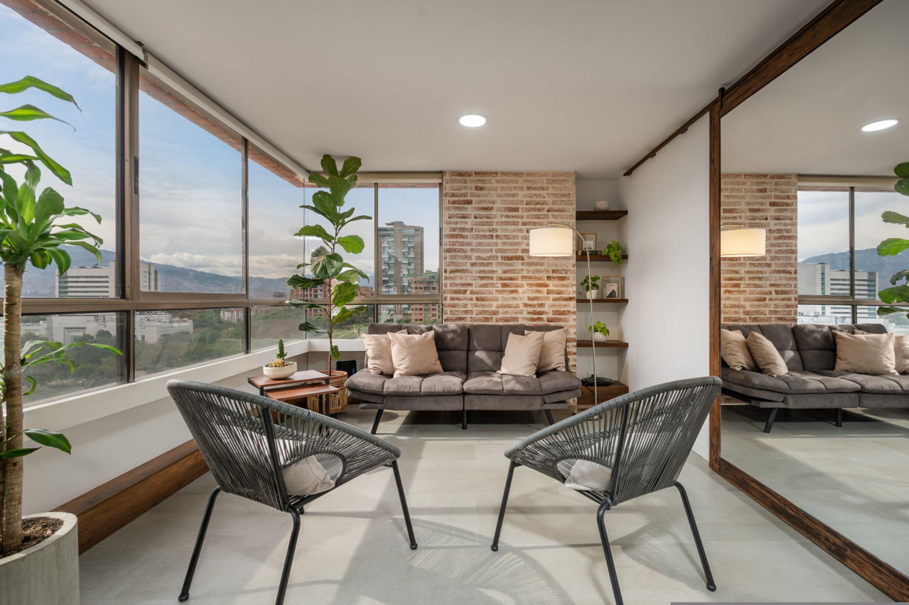 A modern living room with a grey sofa, two black chairs, a brick accent wall, large windows, and potted plants.