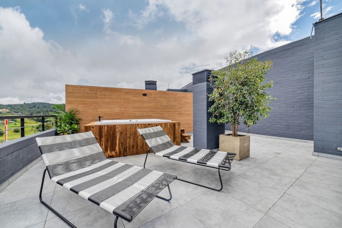 A rooftop patio with two lounge chairs, a wooden table, and a potted plant.
