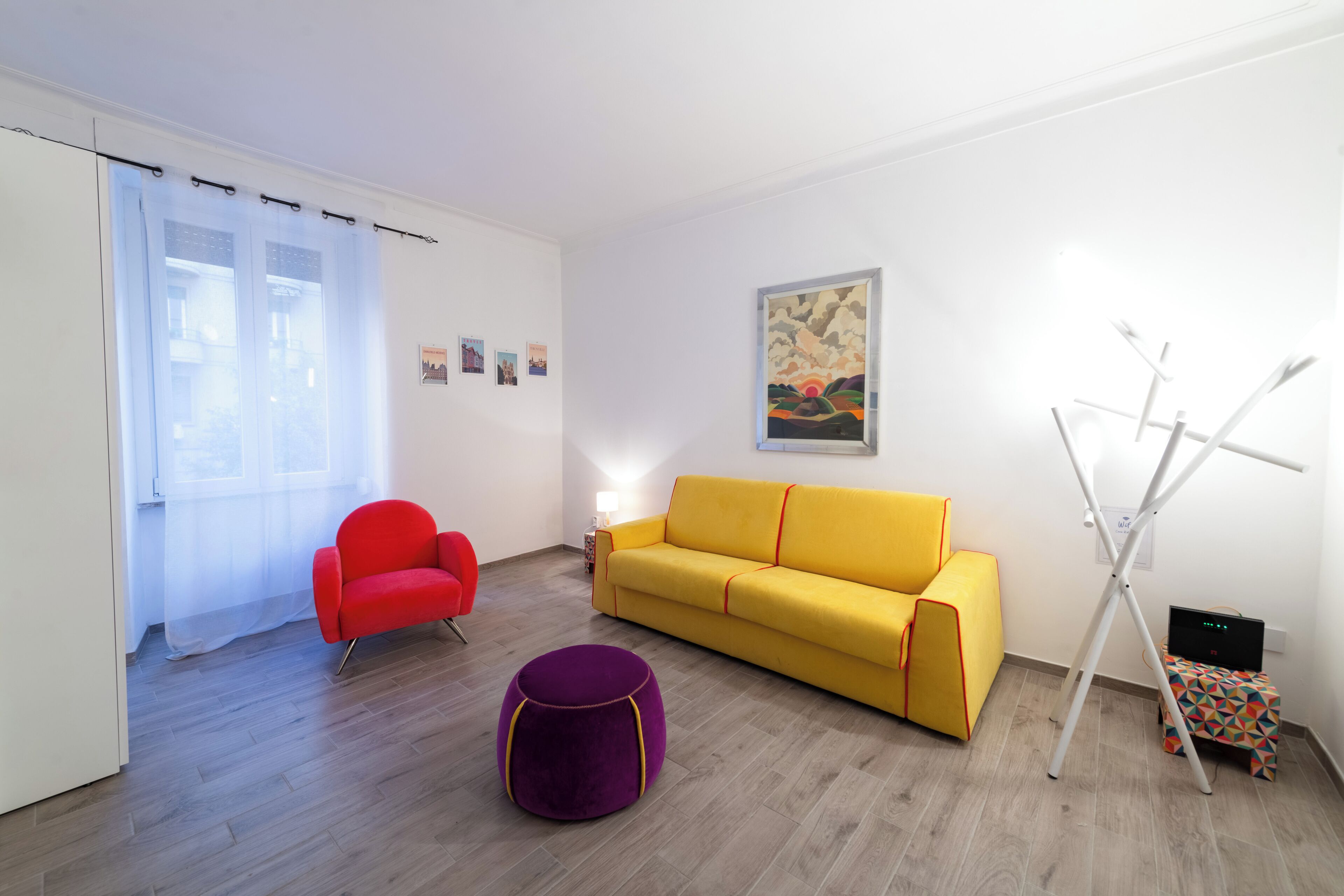 Una habitación con un sofá amarillo, un sillón rojo, un reposapiés morado, un trípode blanco y una pequeña mesa con mantel de estampado geométrico.