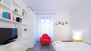 Quarto de hotel moderno com televisão, uma poltrona vermelha, cama e janela com cortina.