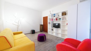 Uma sala de estar moderna com um sofá amarelo, uma poltrona vermelha e um banquinho roxo. Há uma estante branca com livros e uma televisão.