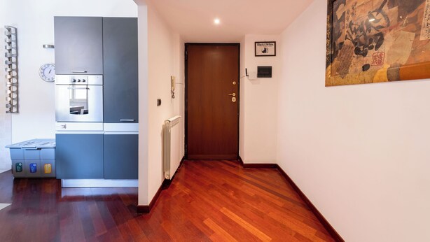 A hallway with a wooden floor, a kitchen area with modern appliances, and a door with a sign above it. - Casa Aosta (Rome)