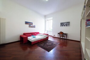 A room with a red sofa bed, a small wooden table, a chair, a patterned rug, and two framed pictures on the walls. - Casa Aosta (Rome)