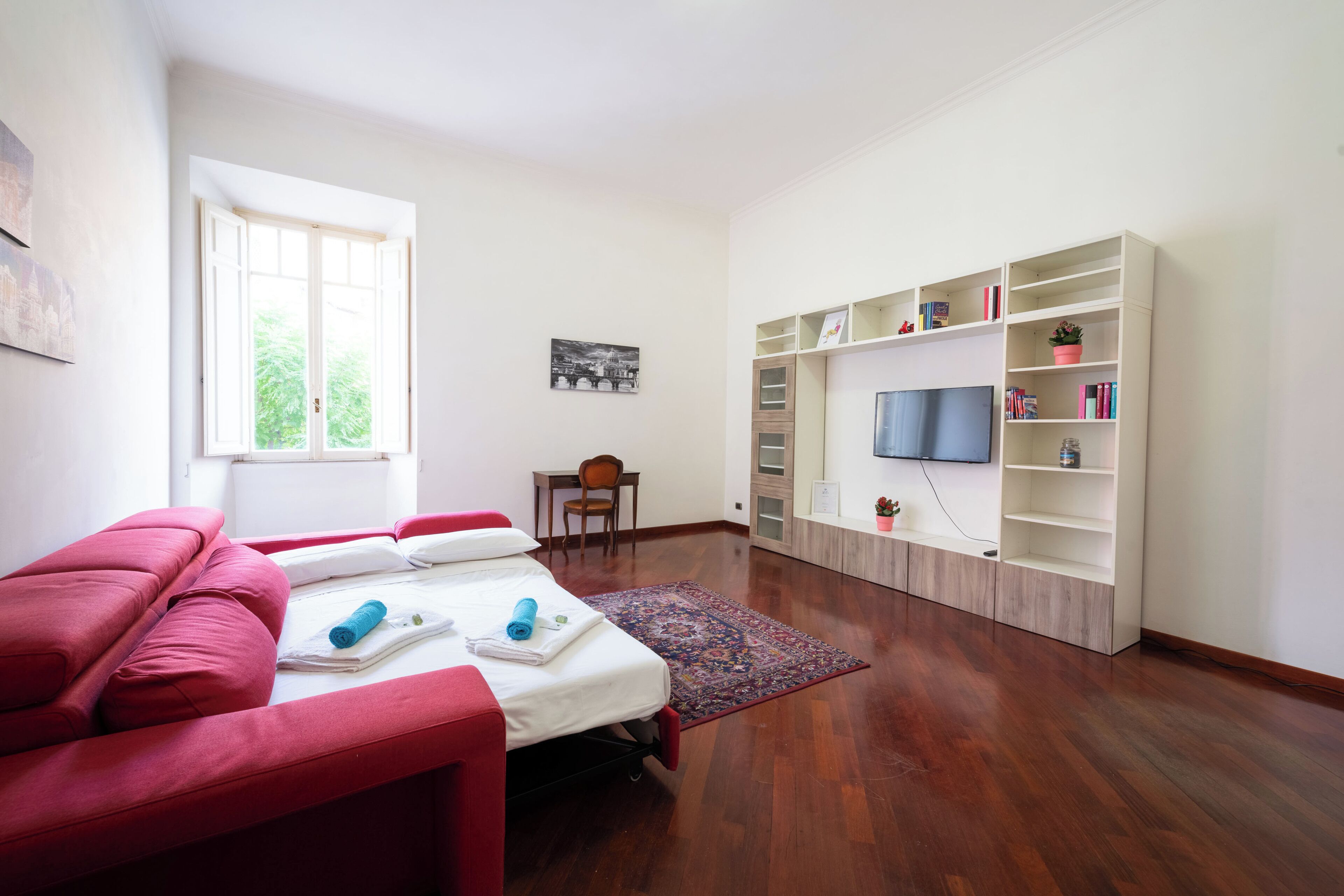 A modern room with a red sofa, a bed with a blue towel, a wooden floor, a bookshelf, and a television.