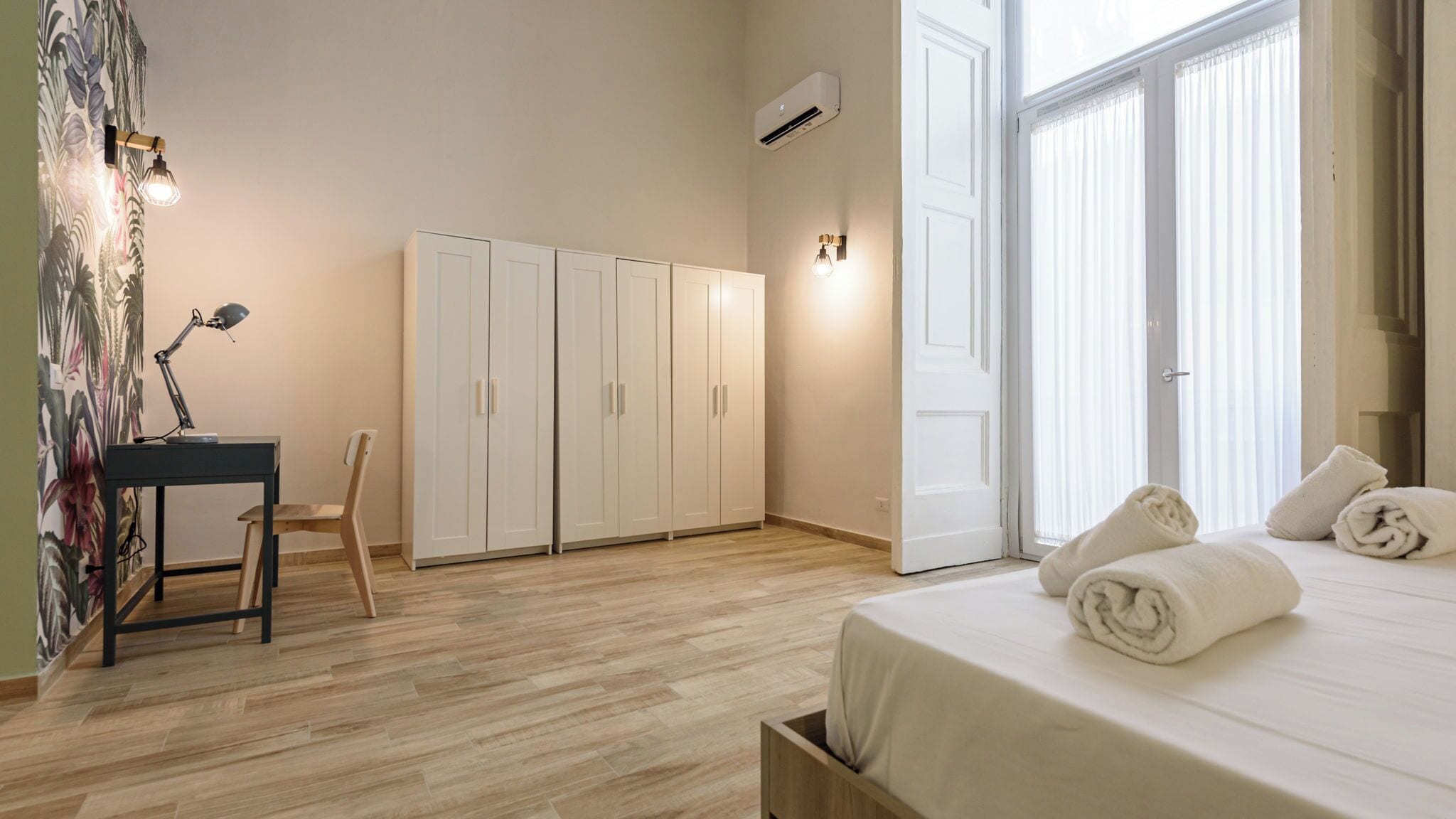 A bedroom with a bed, a desk with a chair, a wardrobe, and a door with curtains.