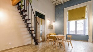 Una habitación con escalera de madera, zona de comedor con mesa y sillas, ventana grande con persiana amarilla, y lámpara fijada a la pared.