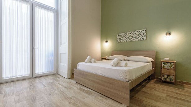 Un dormitorio con cama, mesita de noche, lámpara de pared y ventana con persianas.