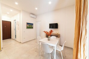 A dining area with a white table, four chairs, a vase with dried plants, a flat-screen TV on the wall, and an air conditioning unit. - Vico Cangiani al Mercato, 20 (Naples)