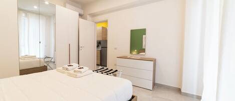 A modern bedroom with a large bed, a wardrobe, a mirror, and a small kitchenette.