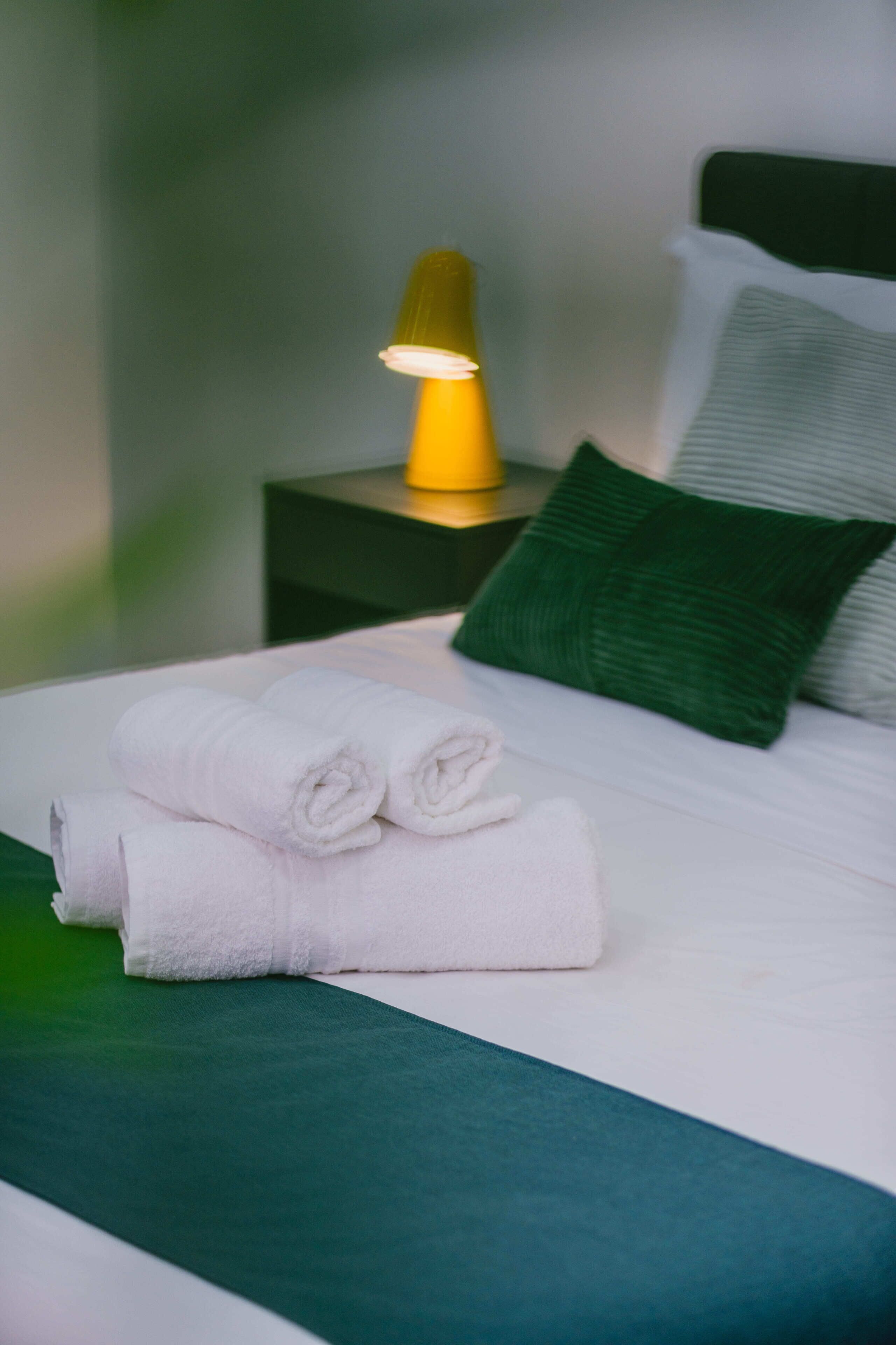 A neatly made bed with white linens and a green throw blanket, a bedside lamp, and a green pillow on a nightstand.
