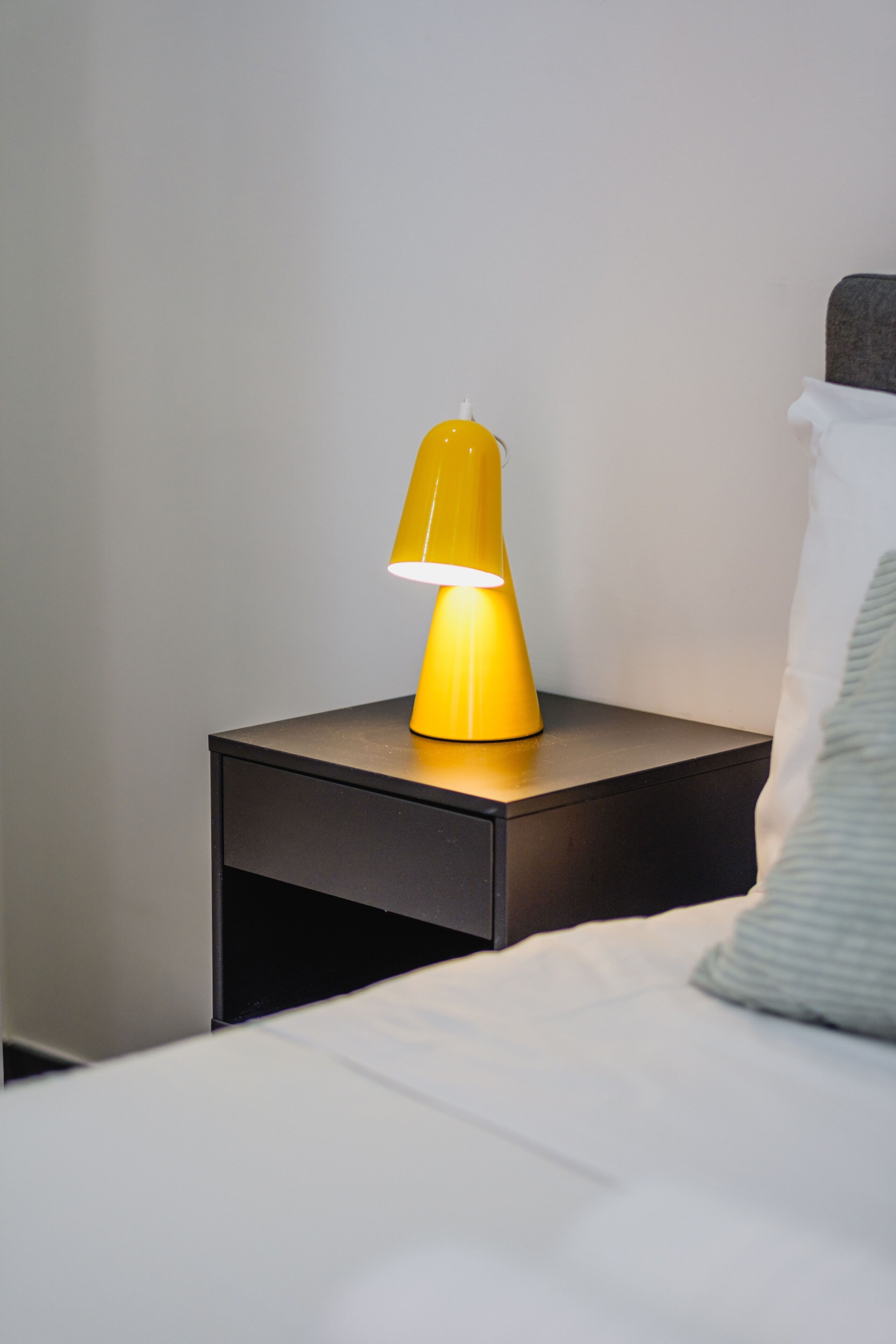 Une table de chevet avec une lampe jaune, un oreiller blanc et une tête de lit grise.