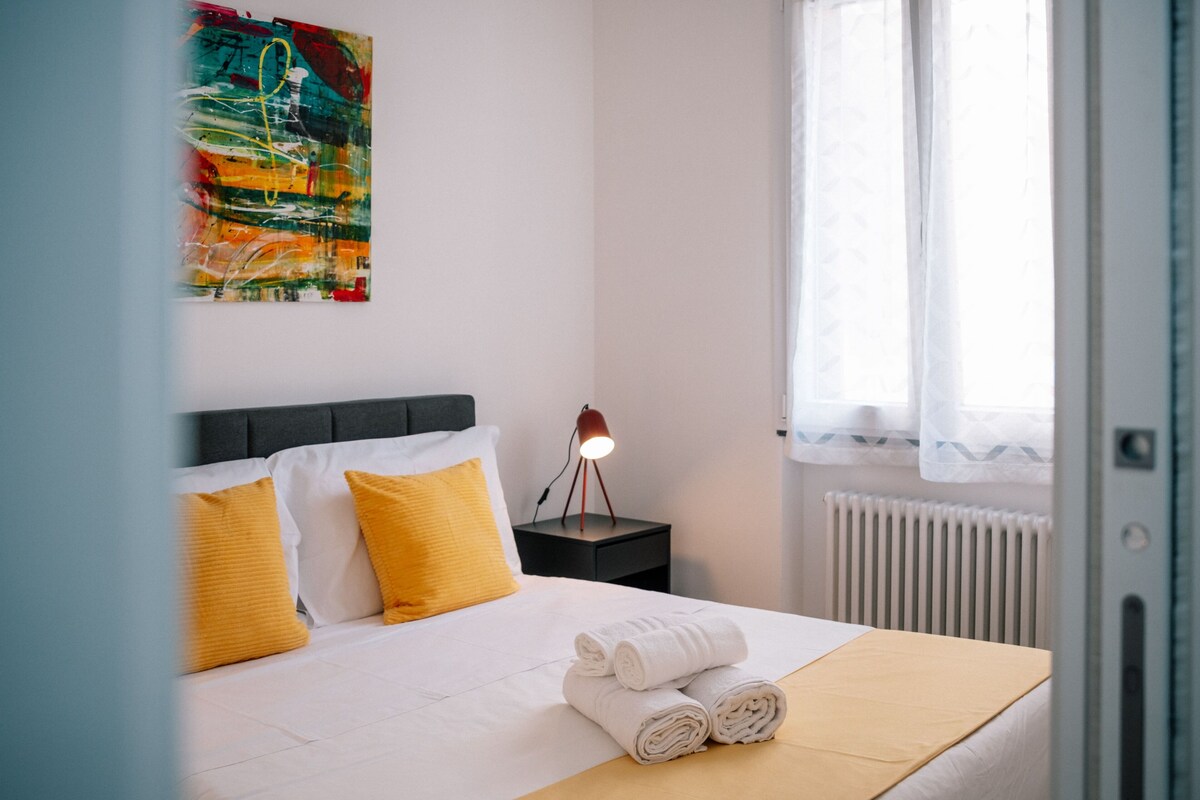 Ein ordentlich bezogenes Bett mit weißen und gelben Kissen, ein aufgerolltes Handtuch, eine Nachttischlampe und eine Heizung.