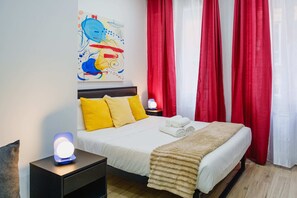 Ein ordentlich bezogenes Bett mit aufgerollten Handtüchern, einer Nachttischlampe und einem Bild an der Wand.