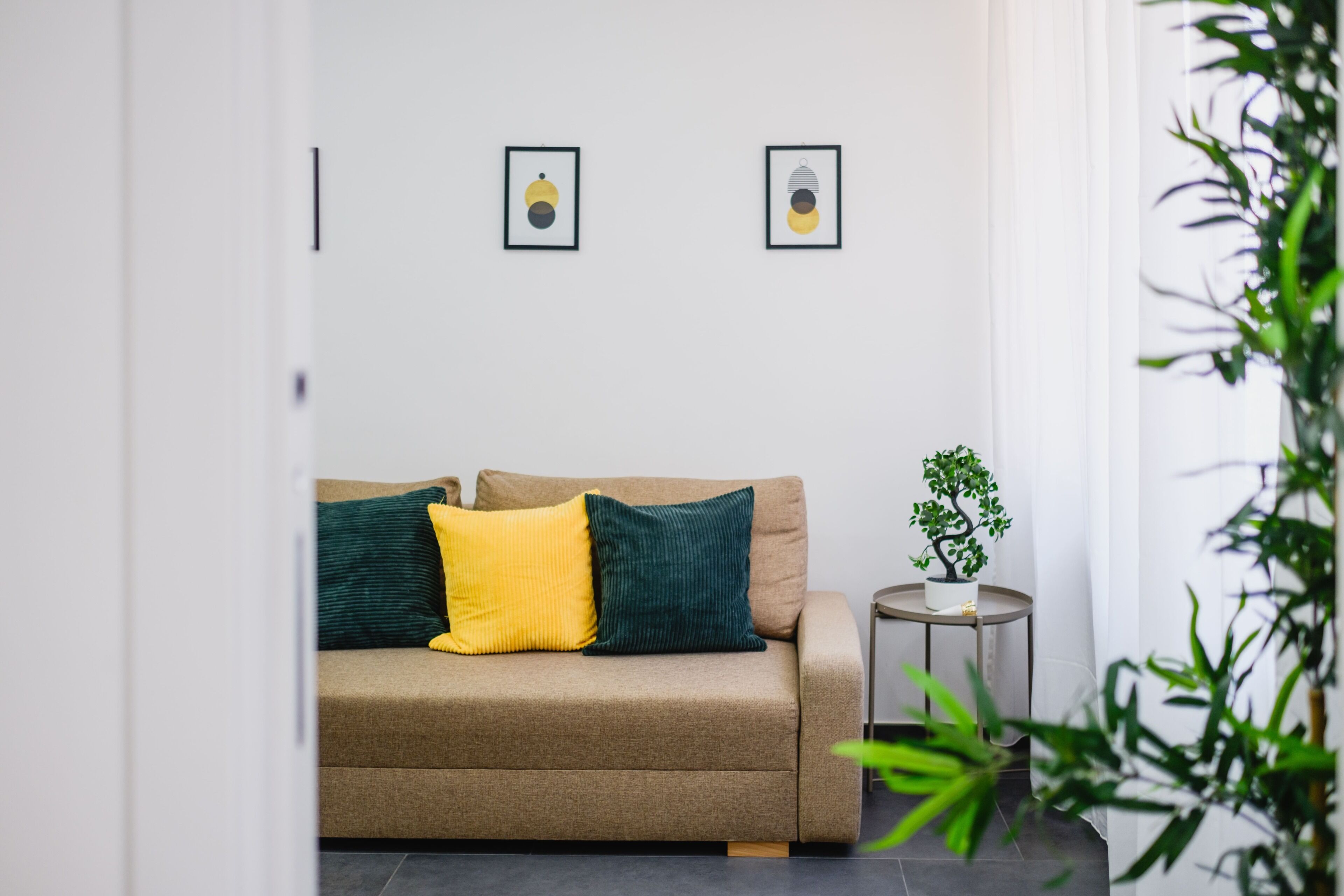 Ein neutral gestalteter Raum mit einer Couch, zwei Kissen, einem kleinen Tisch mit einer Pflanze und zwei gerahmten Bildern an der Wand.