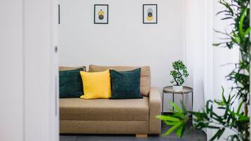 Ein neutral gestalteter Raum mit einer Couch, zwei Kissen, einem kleinen Tisch mit einer Pflanze und zwei gerahmten Bildern an der Wand.