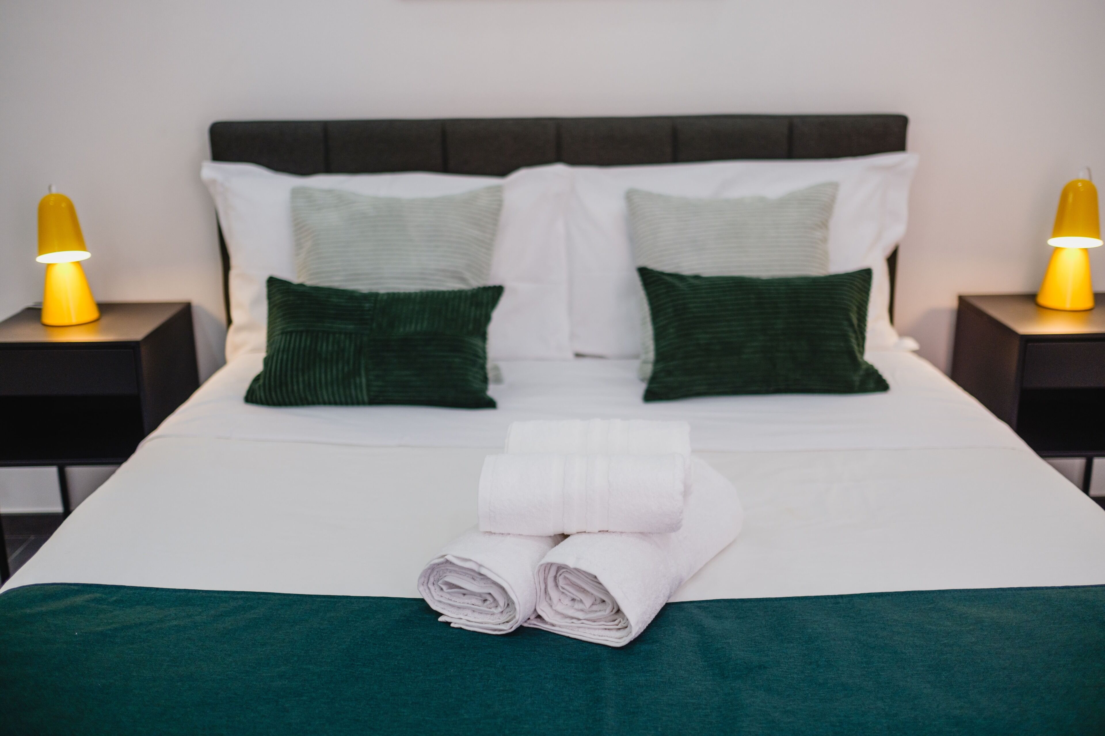 A neatly made bed with white linens and green pillows, two bedside lamps, and a folded towel on the bed.