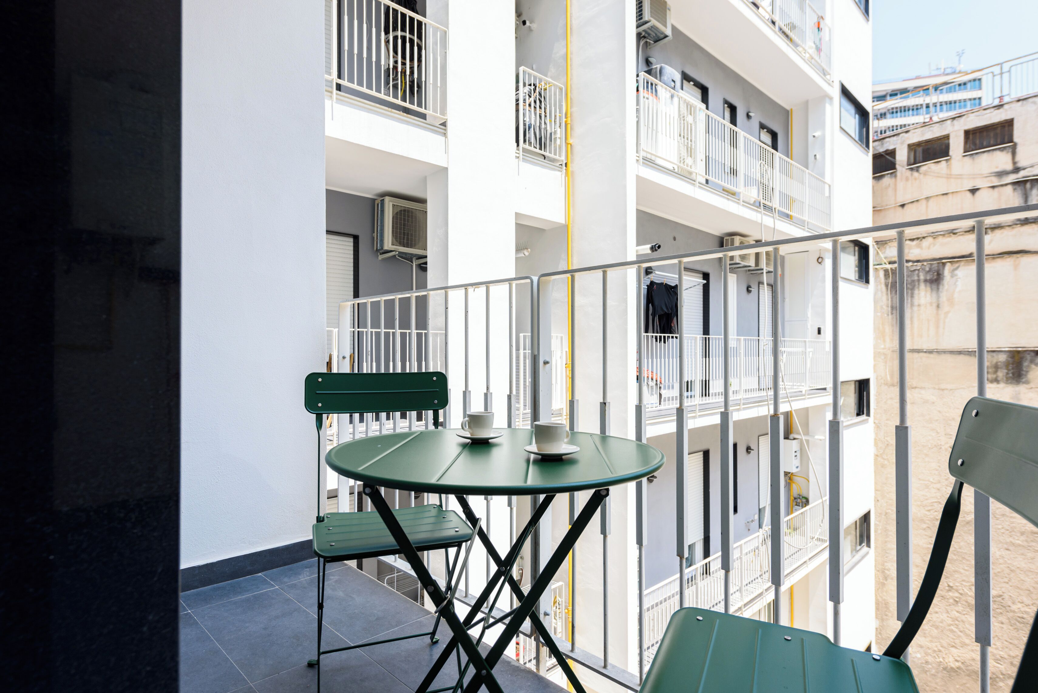 A balcony with a glass table and chairs, overlooking a building with multiple balconies and railings.