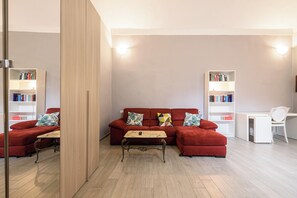 A modern living room with a red sofa, a wooden coffee table, and a white desk with a chair. There are two bookshelves filled with books. - Via Atri 37 (Naples)