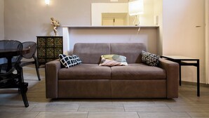 A living room with a brown sofa, two patterned pillows, a glass coffee table, a wooden cabinet with floral designs, and a dining area with a table and chairs. - Via Atri 37 (Naples)