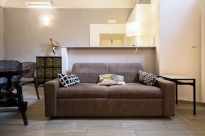Ein Wohnzimmer mit braunem Sofa, zwei gemusterten Kissen, einem Glastisch, einer Holzkommode mit floralen Verzierungen und einem Essbereich mit Tisch und Stühlen.