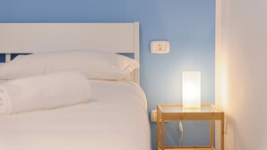 Ein ordentlich bezogenes Bett mit weißen Leinen und einem zusammengerollten Handtuch, ein Nachttisch mit Lampe und ein Lichtschalter an der Wand.