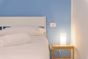 A neatly made bed with white linens and a rolled towel, a bedside table with a lamp, and a wall switch. - Via Aniello Falcone 72b (Naples)