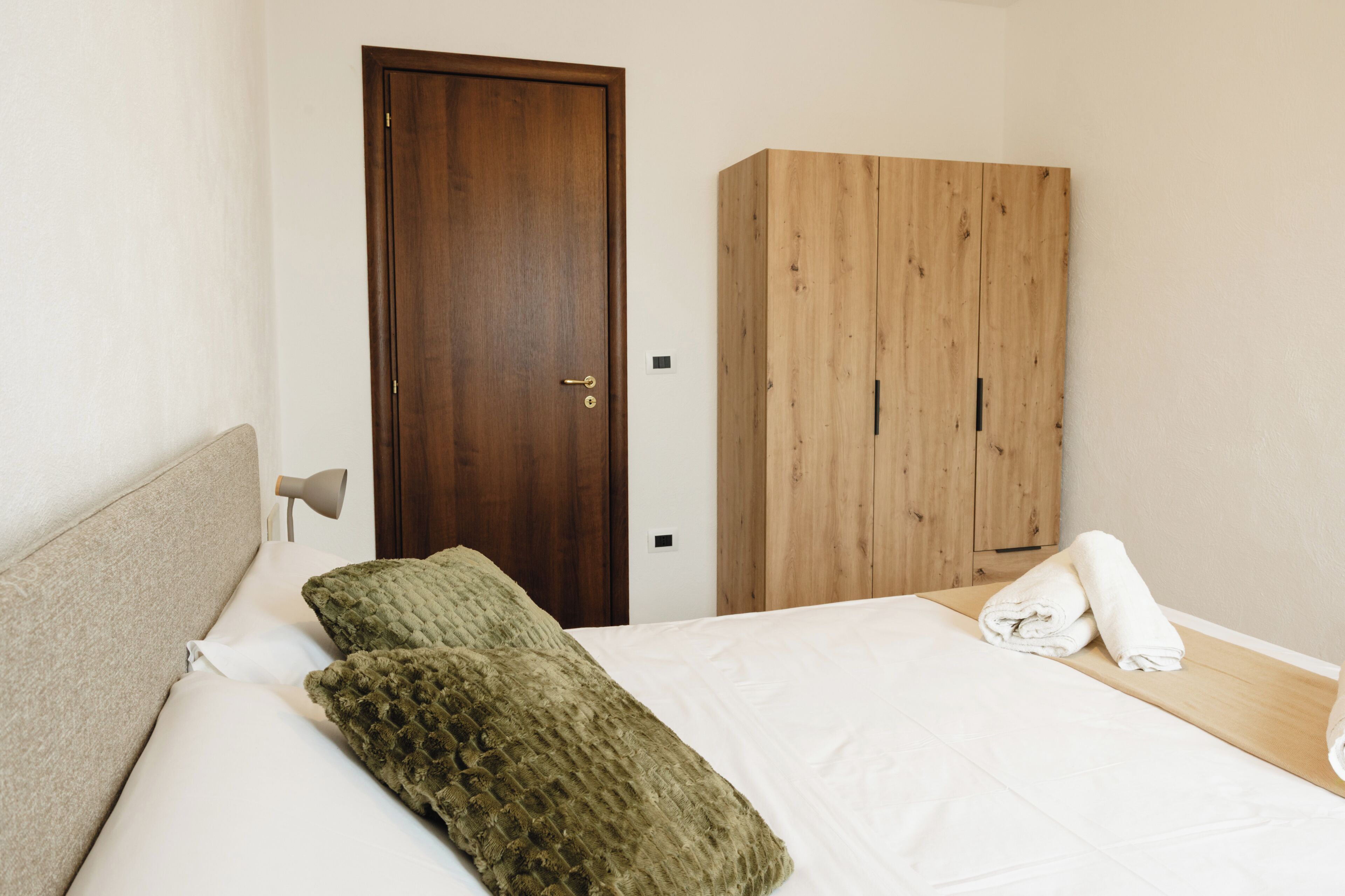 A bedroom with a bed, a wooden wardrobe, a door, and a nightstand with a lamp.