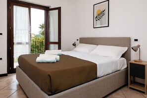 Un dormitorio con cama, mesita de noche, lámpara, un cuadro enmarcado y una puerta corredera de vidrio a un balcón exterior.