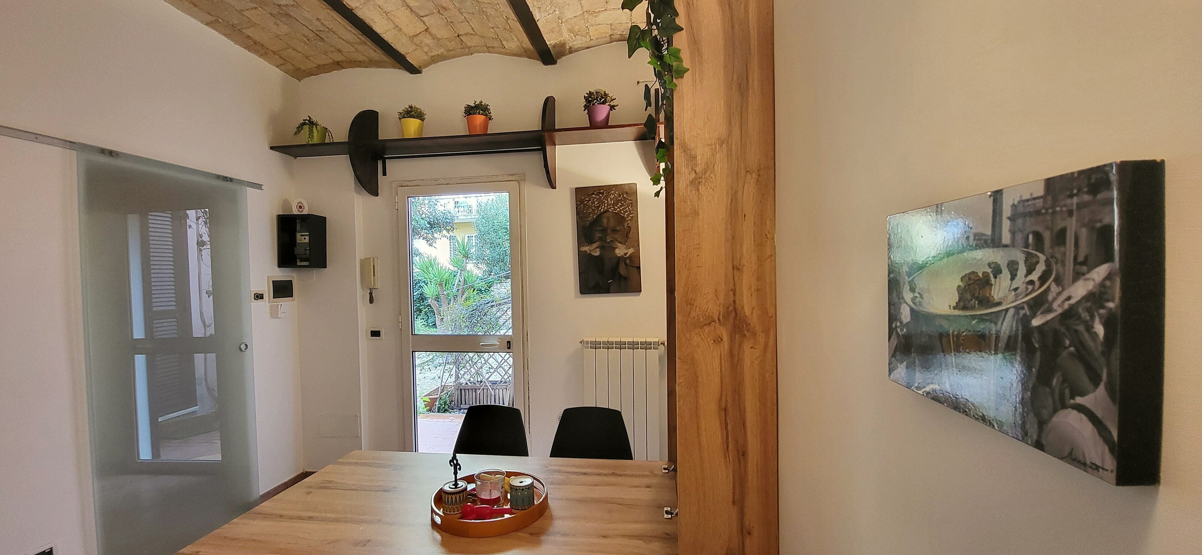 Un coin repas avec une table en bois, des chaises, un plateau contenant une théière, des tasses et un objet décoratif. Il y a une porte sur la gauche, une fenêtre donnant sur un paysage verdoyant et un mur orné d’un tableau encadré.